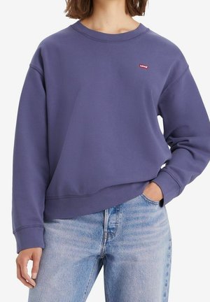Femme portant un sweat-shirt Levi's violet avec un petit logo rouge, assorti à un jean bleu clair, main dans la poche, debout devant un fond blanc.