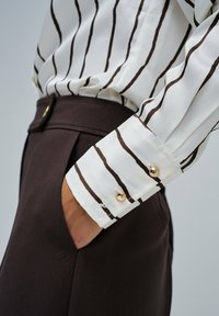 Blusa blanca con rayas verticales marrón, que presenta un puño con botones dorados, combinada con pantalones marrón oscuro de tiro alto. Textura suave.