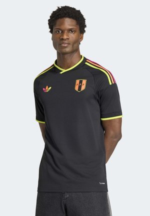 Mann steht und trägt ein schwarzes Sporttrikot mit orange-gelb gestreiftem Logo und mehrfarbigem Adidas-Emblem, neutraler Hintergrund.
