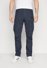 Pantaloni cargo de culoare albastru închis, cu multiple buzunare, detalii cu fermoar și o croială îngustă. Materialul pare a fi durabil și are o textură netedă.