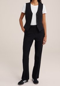 Czarna kamizelka z pięcioma guzikami i dwoma kieszeniami, w połączeniu z czarnymi spodniami. Biała koszulka pod spodem, czarne trampki z trzema białymi paskami.