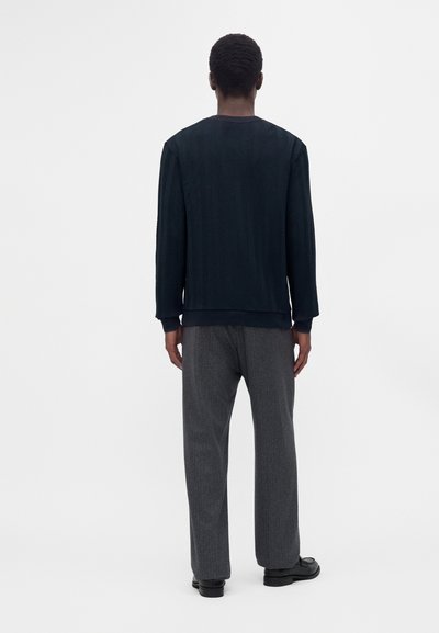 Homme vu de dos, portant un pull texturé foncé, un pantalon gris et des chaussures noires sur un fond clair uni.