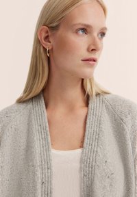 Helle graue Strickjacke mit einem strukturierten Muster, geripptem Kragen und offenem Frontdesign. Trägt sie über einem weißen Oberteil; das Model hat glattes blondes Haar.