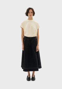 Beige T-shirt met korte mouwen en een klein zakdetail, gecombineerd met een lange zwarte rok met een trekkoord in de taille en witte stiksel.