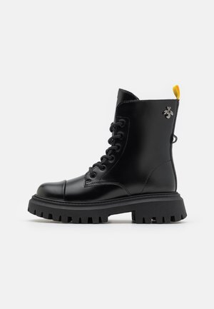 Botte de combat en cuir noir à lacets, semelle épaisse crantée, languette de talon jaune, et petit ornement métallique en forme d’abeille près de la cheville.