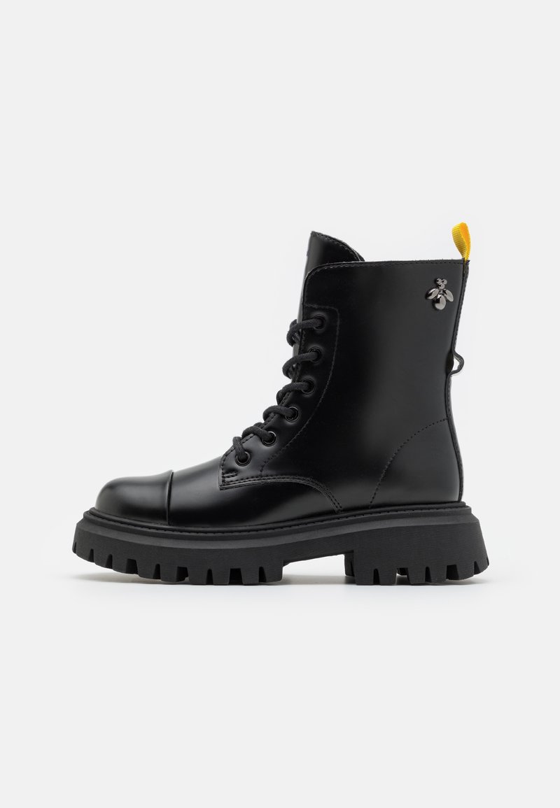 Botte de combat en cuir noir à lacets, semelle épaisse crantée, languette de talon jaune, et petit ornement métallique en forme d’abeille près de la cheville.