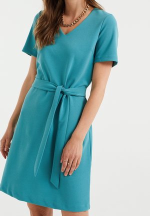 Kvinde iført teal kortærmet kjole med V-hals og matchende stofbælte bundet i taljen, accessorized med en guldkæde halskæde.