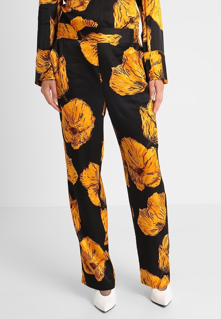Pantalon noir avec un grand motif floral jaune, en tissu léger. Coupe droite, porté avec des talons blancs.