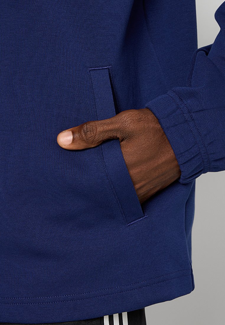 Marineblaue Sweatshirt mit Seitentasche, strukturiertem Stoff, schmalen Bündchen und einem klaren, einfachen Design. Hand ruht auf der Tasche.