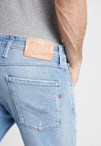 Ljusblå denimjeans med en läderetikett som läser "REPLAY" på midjebandet. Funktioner inkluderar detaljerade bakfickor och blekt textur.