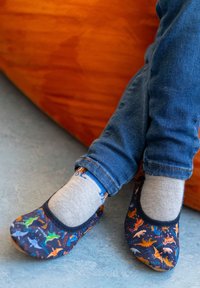 Weiche Stoff-Slipper mit einem dunkelblauen Hintergrund, der farbenfrohe Dinosauriermuster zeigt. Getragen mit grauen Socken und blauen Jeans.