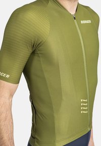 Maillot de cyclisme vert olive avec un design ajusté, manches courtes, texture côtelée sur les épaules, fermeture éclair à l'avant et marquage subtil.