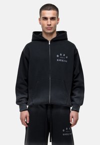 Haut de survêtement zippé noir en coton avec une poche avant, arborant un logo imprimé en blanc. Le design comprend une capuche et des poignets côtelés.