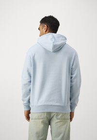 Svetlo modra kapuca s sweatshirtom s mehko teksturo, sproščenim krojem in rebrastimi manšetami ter pasom, nosi se z svetlimi jeans hlačami.
