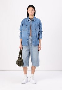 Jaqueta de denim azul claro com dois bolsos no peito e botões, combinada com calções azul claro até ao joelho e ténis brancos. Bolsa verde-oliva.