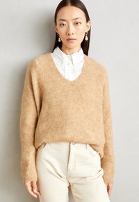 Beige strikket genser med dyp V-hals og ribbestruktur, båret over en hvit skjorte med krage. Designet har løse, lange ermer og en avslappet passform.