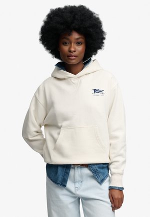 Kvinde med krøllet afro-hår iført en hvid hættetrøje over en blå denimskjorte og lyseblå jeans, stående med den ene hånd bag ryggen.