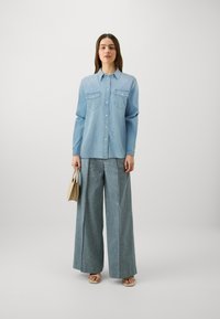 Svijetloplavi traper košulja s dugim rukavima i kopčanjem na gumbe, s dva džepa na prsima, u kombinaciji s širokim svijetloplavim trapericama i ravnim sandalama, drži bež torbu.