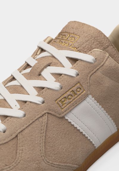 Bež športni čevelj iz semiša z belo usnjeno črto, belimi vezalkami ter zlatim napisom "Polo Ralph Lauren" na jezičku in strani.