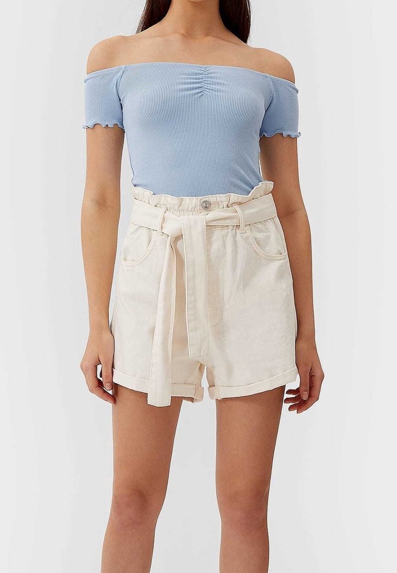 Femme portant un haut côtelé bleu clair épaules dénudées et un short paperbag crème taille haute avec une ceinture nouée.