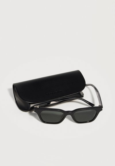 Μαύρα ορθογώνια γυαλιά ηλίου με σκούρους φακούς που ξεκουράζονται μπροστά από μαύρη δερμάτινη θήκη σε ουδέτερο φόντο.