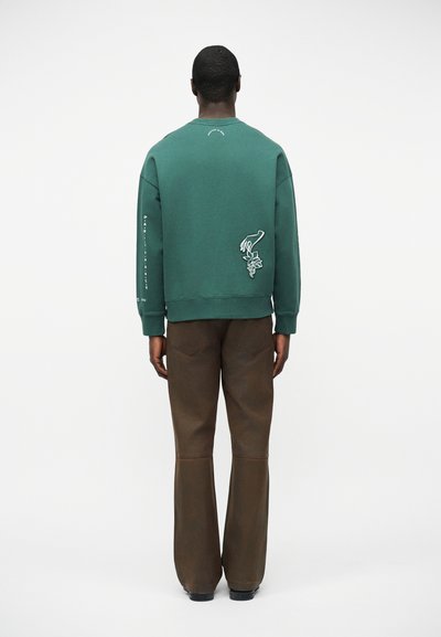 Άνδρας με πλάτη, φοράει πράσινο πουλόβερ με γραφικό χέρι και λουλούδι στην πλάτη και καφέ παντελόνι σε λευκό φόντο.