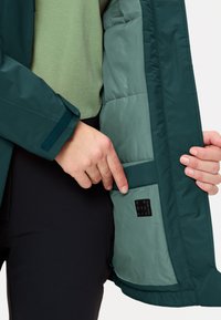 Giacca verde scuro con finitura liscia e opaca, con un interno verde chiaro morbido e una toppa visibile etichettata "RECYCLING". Mano che infila nella tasca interna.