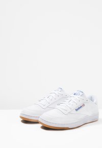 Adidași din piele albă cu vârful rotund, talpă din cauciuc texturat și accente de branding albastre. Prezintă perforații pentru respirabilitate și șireturi.