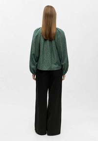 Persoon met lang, steil haar die met de rug naar de camera staat, gekleed in een groene lange mouwen top en losse zwarte broek tegen een effen achtergrond.