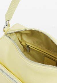 Jasnożółta torebka na ramię z spiralnym uchwytem, otwartym suwakiem oraz widoczną wewnętrzną kieszonką na zamek i podszewką z materiału.