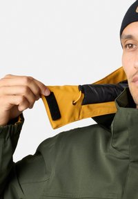 Grön vattentät jacka med en gul inre krage. Har elastiska manschetter, en svart foder och en Velcro-lapp för stängning.