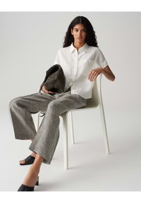 Witte korte mouwen button-up shirt, grijze wijde broek, zwarte hoge hakken sandaaltjes en een gestructureerde donkere tas, zittend in een minimalistische stoel.