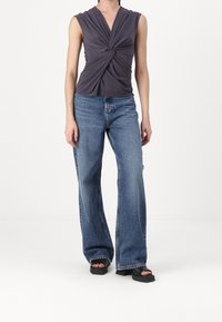 Haut sans manches gris foncé à l'avant tordu, associé à un jean bleu taille haute à jambes larges et à des sandales à talons blocs ouverts noires.