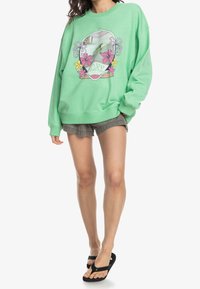Sudadera de gran tamaño en verde menta con un diseño gráfico floral. Combinada con pantalones cortos grises y chanclas negras. Tejido suave, ajuste relajado.