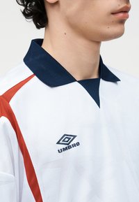 Prim-plan al unei persoane care poartă un tricou sport alb cu dungi roșii, guler naval și logo Umbro naval pe piept.