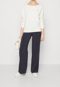 Pull blanc à manches longues avec un design épaules dénudées, associé à un pantalon large bleu marine foncé. Accessoires : un petit sac à main beige et des chaussures blanches.