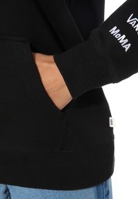 Hoodie noir avec une poche frontale, des poignets côtelés et une étiquette Vans à l'ourlet. Présente un texte sur la manche : "Vans MoMA" en blanc.