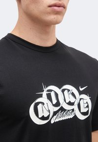 Κοντινό πλάνο ενός ατόμου που φοράει μαύρο t-shirt Nike με λευκά κυκλικά γράμματα που σχηματίζουν τη φράση «Nike Athletics» στο στήθος.