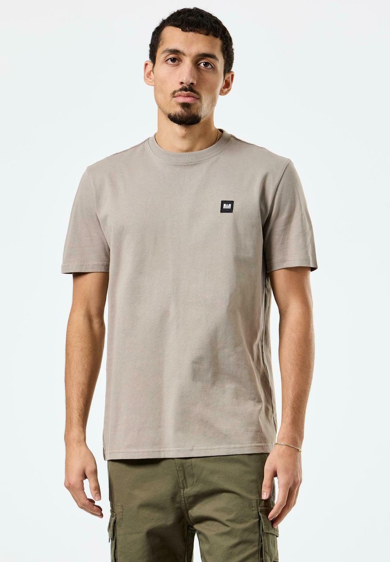 Lys beige t-shirt med korte ærmer i bomuld, med et lille sort firkantet mærke på brystet. Rund halsudskæring, afslappet pasform.