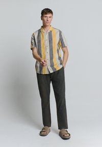 Korte mouwen overhemd met knopen in geel, grijs en witte strepen, gecombineerd met donkere broek en bruine sandalen. Heeft een kraag.