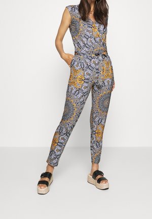 Woman wearing sleeveless, patterned jumpsuit with yellow, black, and gray details, paired with black platform sandals.