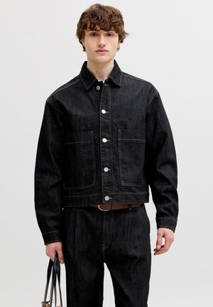 Junger Mann, der eine schwarze Jeansjacke und schwarze Jeanshose trägt und eine schwarze Tragetasche vor einem einfarbigen hellen Hintergrund hält.