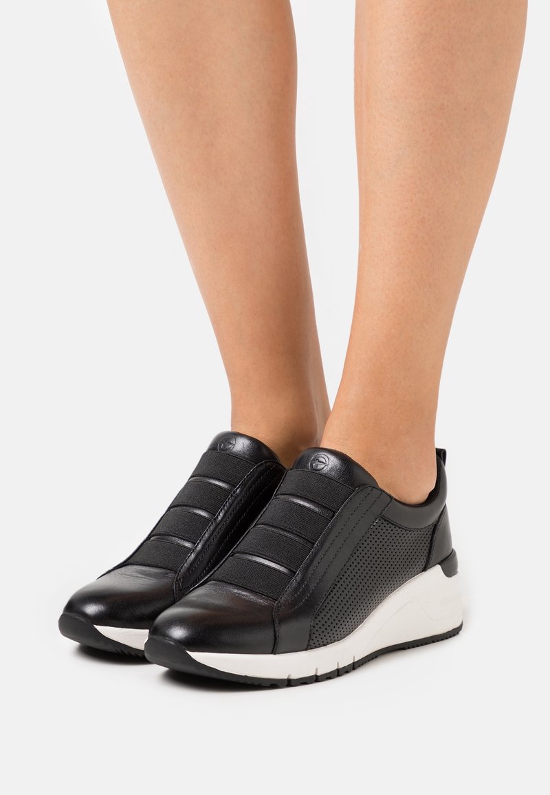 Pieds portant des baskets compensées noires à enfiler avec fermetures à bandes élastiques et semelles blanches sur un fond clair.