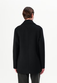 Zwarte wollen jas met een gestructureerd ontwerp, voorzien van een geknipt kraag en zichtbare stiksels. De achterkant toont een rechte snit en minimale accenten.