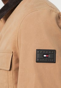 Tan stofjakke med krave i brunt fløjl, der har en sort rektangulær lap med "Tommy Hilfiger" logoet og farveblok emblem.