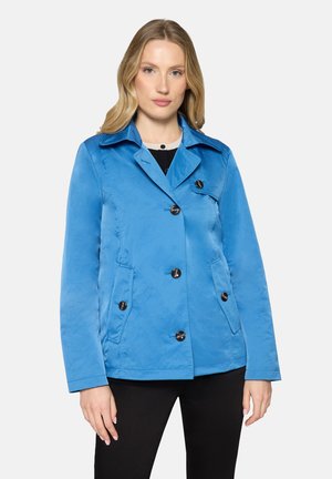 Vrouw die een felblauwe jas met knopen, kraag en voorzakken draagt, samen met een zwarte broek, staand voor een effen witte achtergrond.