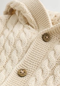 Sweter z dzianiny w kolorze kremowym z wzorem w warkocz, złożonym kapturem i dużymi brązowymi guzikami z wygrawerowanym napisem. Widoczne detale tekstury materiału.
