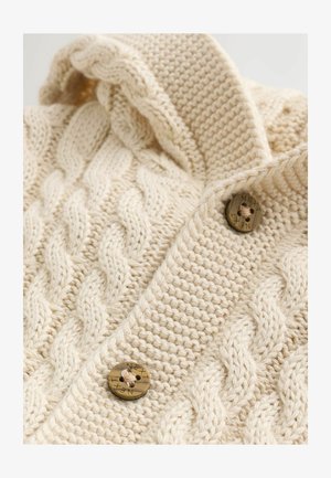 Krémový pletený svetr s kabelovým vzorem, se skládacím kapucí a velkými hnědými knoflíky s vyrytým textem. Viditelné detaily texturované tkaniny.