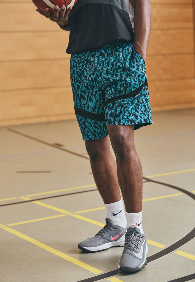 Shorts de basket en motif léopard bleu avec des accents noirs, associés à des chaussures Nike grises et roses et des chaussettes blanches, sur un fond de sol de gym.