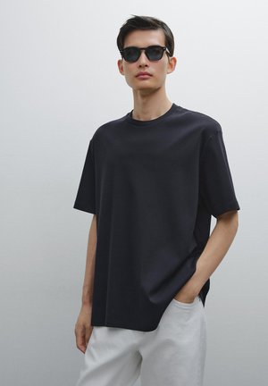 Sort crew-neck t-shirt lavet af glat stof; løs pasform med korte ærmer. Modellen bærer sorte solbriller og lyse bukser.